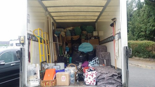 The image shows the interior of an open cargo van, likely used for rubbish removal or transportation, parked outdoors near a residential or urban area with trees and hedges visible in the background. Inside the van, various waste materials and items are stored, including large bags of mixed refuse in dark green and black, some partially covered with a light blue sheet. There are cardboard boxes of different sizes, some closed and others open, containing miscellaneous objects. A yellow hand truck with a handle and wheels is positioned on the left side, leaning against the van’s interior wall. The floor of the van is cluttered with smaller cardboard boxes, a clear plastic container, and patterned fabric or padding. The van's interior walls are metallic, with a slightly reflective surface, and the ceiling is made of ribbed aluminum. The environmental lighting suggests daylight, and the scene indicates typical rubbish collection or cleanup activity, as may be undertaken by Business Waste Removal Anerley in the local area close to postcode SE20.