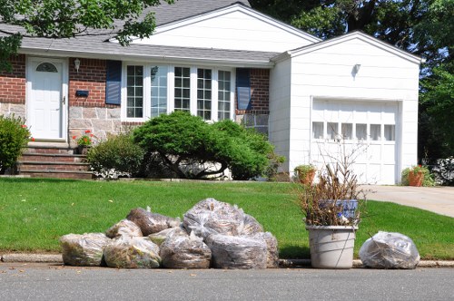 On a residential street in Anerley, a collection of large, irregularly shaped stones with rough textures and earthy tones are arranged along the curbside in front of a neatly maintained front garden. To the right of the stones, a white plastic garden pot contains dried, brown plant stems and twigs, adding a natural element to the scene. Behind this arrangement, a well-kept lawn with vibrant green grass extends towards a brick and white-panelled house with a large bay window and a white front door, slightly elevated by a small set of brick steps. A driveway leading to an attached garage with a white garage door is visible on the right side of the house. The overall scene captures an everyday moment of outdoor clearing in a suburban area, with the stones and plant debris awaiting removal, consistent with rubbish removal services carried out by Business Waste Removal Anerley in the local SE20 postcode area. The natural lighting suggests a bright day, emphasizing the clear, tidy appearance of the property and street.