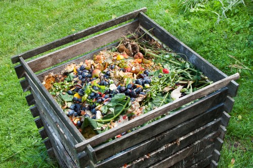 A black wooden crate with horizontal slats is positioned on a grassy area, filled with a mixture of garden waste, including fallen leaves, small branches, vegetable scraps, and flowering plant stems. The surface of the waste appears damp and partially decomposed, with various textures from smooth leaves to rougher, woody stems. The crate is situated outdoors, with a background of green grass and some plants, indicating a garden or backyard setting. The overall scene suggests the collection of garden refuse prior to removal or composting, consistent with rubbish removal services in Anerley. The wooden slats of the crate show slight weathering, and the waste inside is disorganized, highlighting typical garden clearance activity that a professional waste removal company such as Business Waste Removal Anerley might assist with.