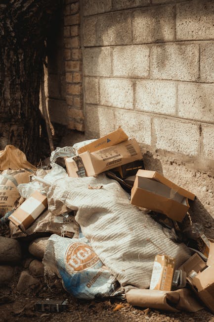 Two large black rubbish bags, made of plastic with a glossy surface, are placed on a paved sidewalk near a black metal fence with vertical bars and a wooden lattice panel behind it. The bags appear to
